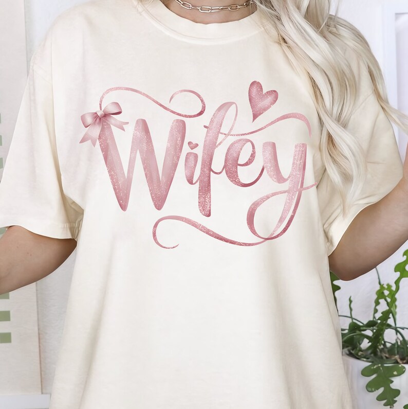 May include: A white t-shirt with the word "Wifey" written in pink glitter lettering with a pink bow and a pink heart.