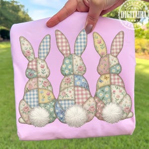 May include: A light pink sweatshirt featuring three patchwork bunny designs. Each bunny has a fluffy white tail and is decorated with floral and gingham patterns in pastel colors. The design is held by a person's hand.