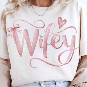 May include: A white t-shirt with the word "Wifey" written in pink glitter font. The word is surrounded by a pink glitter swirl design. A pink glitter bow is on the left side of the word and a pink glitter heart is on the right side of the word.