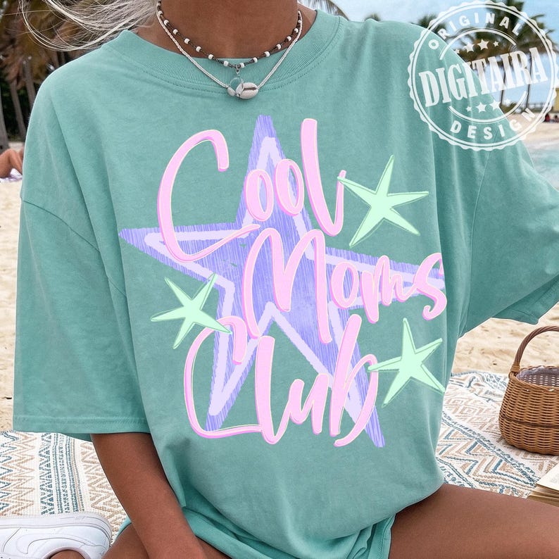 May include: A light teal t-shirt with the text "Cool Moms Club" in pink script over a purple star and green stars. The shirt is a casual style, perfect for everyday wear.