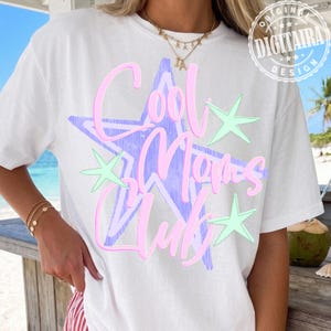 May include: White t-shirt with a large, pastel design. The design features a blue star shape with the words "Cool Moms Club" in pink script. Accenting the star are several light green star shapes. A gold necklace and bracelets are visible.