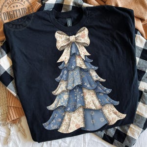 Peut inclure: Un t-shirt bleu marine avec un motif d'arbre de Noël. L'arbre est fait de tissu floral bleu et crème, avec un grand nœud en haut. Le t-shirt est de la marque Gildan.