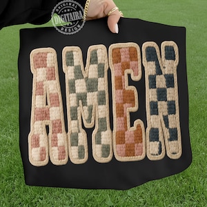 May include: Black tote bag with the word "AMEN" in large, textured letters. The letters are in a checkerboard pattern of pink, green, brown, and blue, with a beige border. The bag is held against a green grass background.