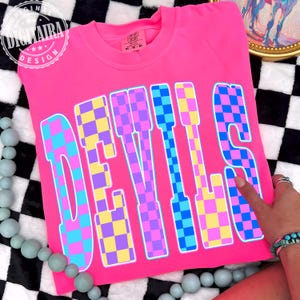 May include: A bright pink t-shirt with the word "DEVILS" in large, checkered letters. The letters are a mix of blue, purple, and yellow squares. The shirt has a crew neck and short sleeves. The brand name "DIGITALRA DESIGN" is in a circle in the upper left corner.