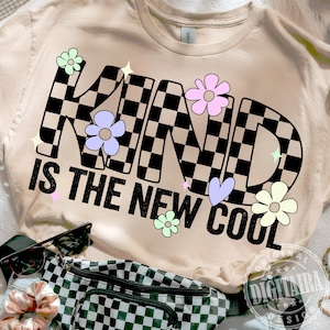 May include: Cream-colored t-shirt with the words "KIND IS THE NEW COOL" in black, with the word "KIND" in a checkered pattern. The design includes flowers, stars, and a heart.