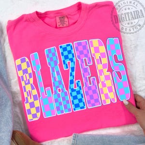 Puede incluir: Una camiseta rosa brillante con la palabra "BLAZERS" en letras grandes a cuadros. El patrón de cuadros incluye cuadrados azules, amarillos y morados. La camiseta tiene cuello redondo y mangas cortas.