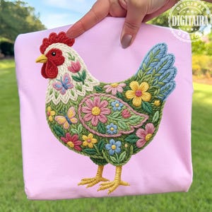 May include: A light pink sweatshirt featuring a detailed, embroidered chicken design. The chicken is adorned with colorful flowers, butterflies, and intricate stitching. The design includes red, yellow, pink, blue, and green hues, creating a vibrant and artistic look.