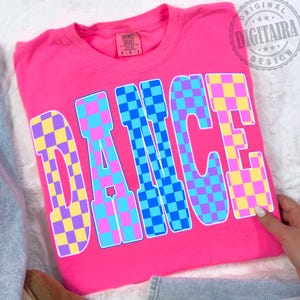 Puede incluir: Una camiseta rosa intenso con la palabra "DANCE" en letras grandes a cuadros. Las letras son una mezcla de cuadrados azules, rosas, morados y amarillos. La camiseta está doblada y parece estar hecha de un material suave.