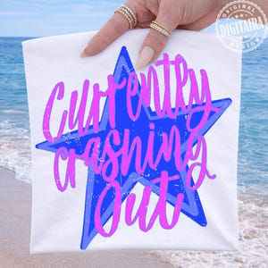 May include: White t-shirt featuring a blue star with the text "Currently Crashing Out" in pink script. The design is held up against a backdrop of the ocean and sky. The shirt is a casual wear item.
