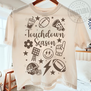 Puede incluir: Camiseta color crema con un diseño temático de fútbol americano. El diseño incluye un balón de fútbol, un casco, un megáfono y el texto "Touchdown Season". Elementos adicionales incluyen estrellas, una flor y un rayo. La camiseta se muestra en una percha de madera.