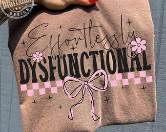 Pode incluir: Uma camiseta marrom com a frase "Effortlessly Dysfunctional" em letras pretas, adornada com flores rosa, um laço e um padrão xadrez rosa e branco. A camiseta é segurada por uma mão.