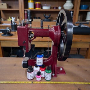 May include: A vintage burgundy sewing machine with a large black flywheel. The machine has the word "CUB" on it. Several bottles of acrylic leather paint in various colors, including black, white, red, pink, blue, and green, are in front of the machine.