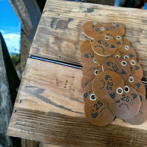 May include: A pile of brown leather tags with the text "Straight 25" printed on them. Each tag has a hole punched in the top and a silver metal ring.