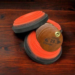 May include: A close-up shot of orange clay targets with black rims and a stack of brown leather tags. The tags have the text "CRUSH 25 STRAIGHT" printed on them. The items are on a dark wood surface.