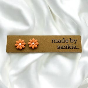 May include: A pair of orange flower-shaped stud earrings with white centres, attached to a rectangular brown display card featuring the black printed text made by saskia. The earrings have eight rounded petals each and a smooth matte finish.
