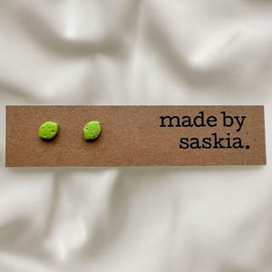 May include: A pair of small, lime green stud earrings with a textured surface, attached to a rectangular brown cardboard display card. The card features the black printed text "made by saskia." positioned to the right of the earrings.