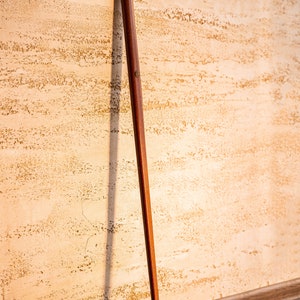 May include: A wooden walking cane with a dark brown shaft and a decorative brass handle. The handle has an ornate design. The cane is leaning against a textured beige wall and a wooden floor.