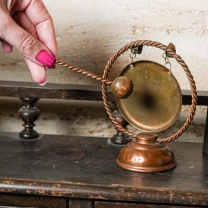 May include: A small, antique-style gong with a copper-colored frame and base. The gong itself is a golden color, and a wooden mallet is held near it. The gong is displayed on a dark wooden surface.