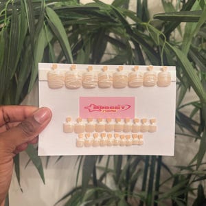 May include: A set of 20 beige square-shaped press-on toe nails with white swirl designs. The nails are displayed on a white card with the text "BUDGET nails" in pink.