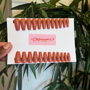 Puede incluir: Un juego de 20 uñas postizas de color nude en forma de ataúd. Las uñas están dispuestas en dos filas en una tarjeta blanca con el texto "BUDGET nails" en rosa.
