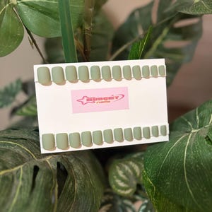 May include: A set of sage green press-on nails arranged on a white card. The card features a pink logo that reads "BUDGET nails". The nails are square-shaped and displayed against a backdrop of lush green foliage.