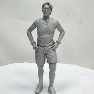 May include: A gray figurine of a person in athletic attire. The figure is wearing a tank top, shorts, and sneakers. The person has their hands on their hips and is standing on a white surface.