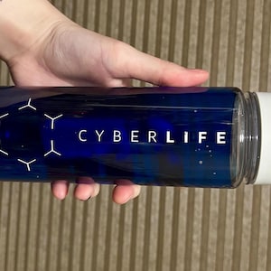Könnte beinhalten: Eine transparente, zylindrische Wasserflasche mit dunkelblauem Innenraum und weißem Deckel. Die Flasche trägt den weißen Schriftzug "CYBERLIFE" und ein geometrisches Design. Die Flasche wird von einer Hand gehalten.