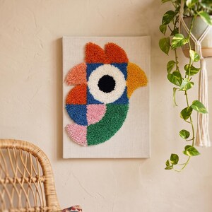 Peut inclure: Une œuvre d'art murale rectangulaire présentant un motif d'oiseau stylisé réalisé en broderie punch needle. L'oiseau est composé de formes géométriques en laine orange, jaune, bleue, rose et verte, avec un grand œil circulaire blanc et noir.