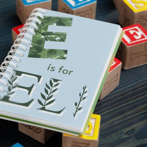 May include: A blue spiral bound book with the letter "E" in green on the cover. The book says "E is for" and has green leaves around the letter. The book is surrounded by wooden alphabet blocks with letters like "S", "U", and "T".