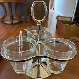 May include: A silver condiment holder with three glass jars and a handle. The holder has a round base and three curved arms that hold the jars.
