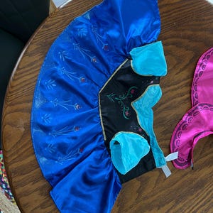 Puede incluir: Un vestido de disfraz azul y negro con ribete dorado. El vestido tiene una falda azul con un dise&ntilde;o floral y un corpi&ntilde;o negro con detalles turquesa. Un accesorio rosa es visible a la derecha.