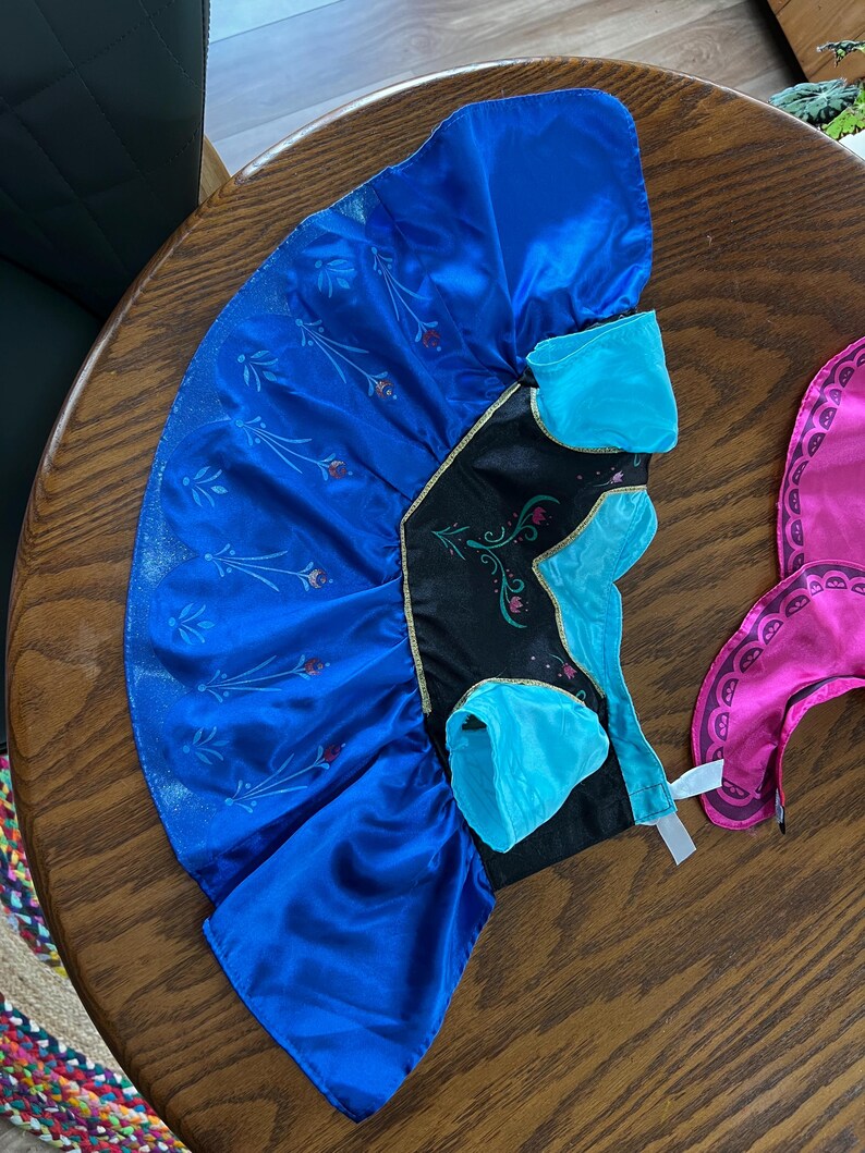 Puede incluir: Un vestido de disfraz azul y negro con ribete dorado. El vestido tiene una falda azul con un dise&ntilde;o floral y un corpi&ntilde;o negro con detalles turquesa. Un accesorio rosa es visible a la derecha.