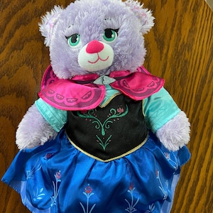 Puede incluir: Un oso de peluche lavanda que lleva un vestido azul y negro con una capa rosa. El oso tiene ojos verdes, una nariz rosa y un hocico blanco. El vestido tiene un dise&ntilde;o floral. El oso est&aacute; sobre una superficie de madera.