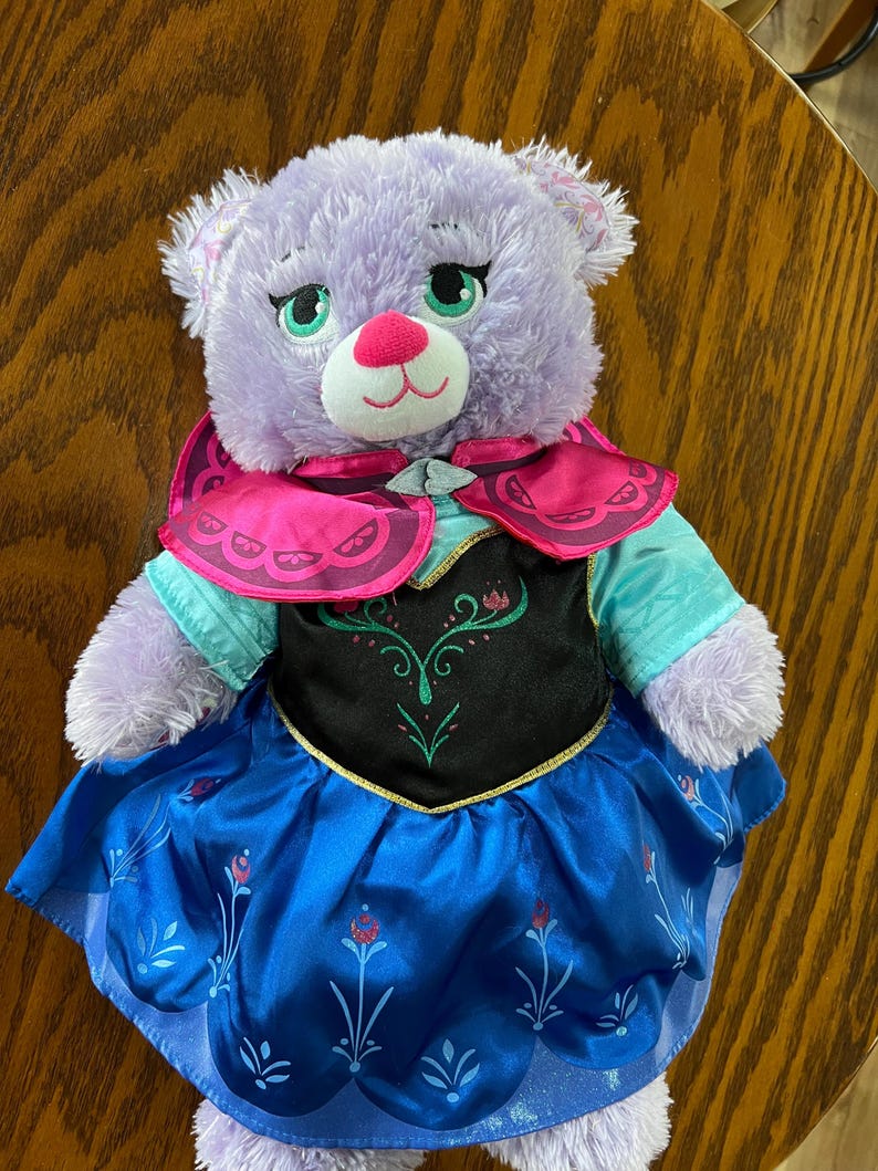 Puede incluir: Un oso de peluche lavanda que lleva un vestido azul y negro con una capa rosa. El oso tiene ojos verdes, una nariz rosa y un hocico blanco. El vestido tiene un dise&ntilde;o floral. El oso est&aacute; sobre una superficie de madera.