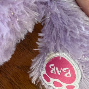 Puede incluir: Primer plano de una pata de peluche lavanda con una almohadilla rosa en forma de coraz&oacute;n. La almohadilla presenta las letras "BAB" en blanco. La piel de felpa tiene una textura suave y esponjosa con acentos de purpurina plateada.