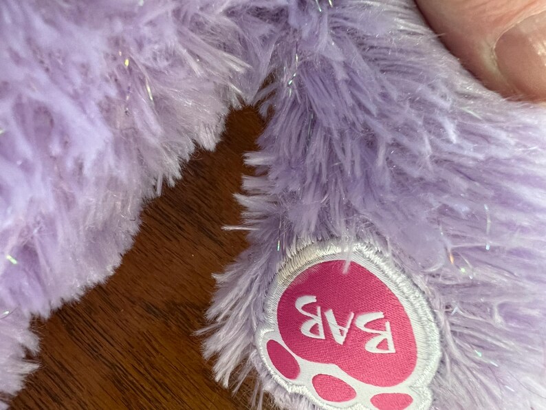 Puede incluir: Primer plano de una pata de peluche lavanda con una almohadilla rosa en forma de coraz&oacute;n. La almohadilla presenta las letras "BAB" en blanco. La piel de felpa tiene una textura suave y esponjosa con acentos de purpurina plateada.