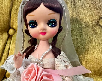 May include: A vintage bride doll with dark brown hair styled in pigtails. The doll wears a white lace dress, a pearl necklace, and a veil. A pink ribbon and two pink fabric roses are at the waist. The doll has large, expressive eyes.