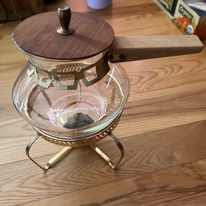 May include: A glass coffee pot with a wooden lid and handle, sitting on a gold metal stand. The pot has a gold trim around the top.