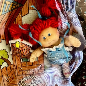 May include: A vintage doll with bright red yarn hair and blue eyes, wearing a blue polka dot dress with white lace trim. The doll is in a cardboard display with a wooden cabin illustration and the word "TEAM".