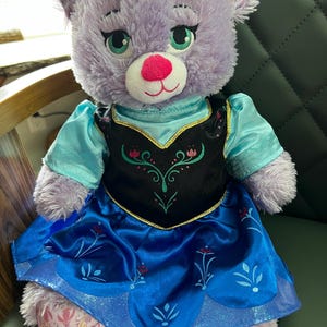 Puede incluir: Un oso de peluche lavanda que lleva un vestido azul y negro con detalles florales. El oso tiene ojos verdes y una nariz rosa. El vestido tiene un ribete dorado. El oso est&aacute; sentado sobre una superficie verde.