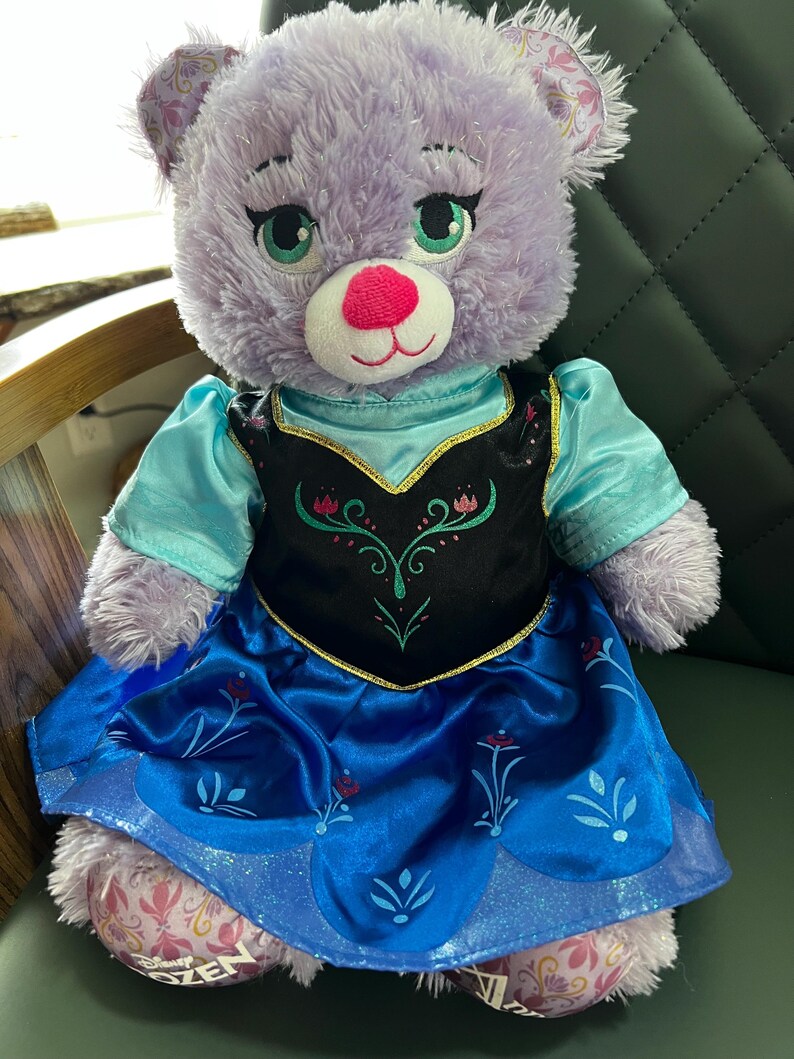 Puede incluir: Un oso de peluche lavanda que lleva un vestido azul y negro con detalles florales. El oso tiene ojos verdes y una nariz rosa. El vestido tiene un ribete dorado. El oso est&aacute; sentado sobre una superficie verde.