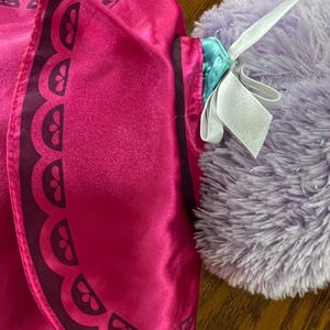 Puede incluir: Primer plano de una capa de disfraz rosa con un borde festoneado marr&oacute;n oscuro. La capa se combina con un peluche morado y esponjoso con una cinta blanca atada alrededor del cuello. La capa tiene una textura satinada.