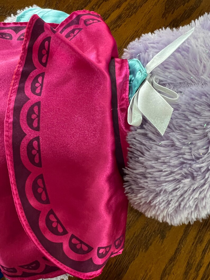 Puede incluir: Primer plano de una capa de disfraz rosa con un borde festoneado marr&oacute;n oscuro. La capa se combina con un peluche morado y esponjoso con una cinta blanca atada alrededor del cuello. La capa tiene una textura satinada.