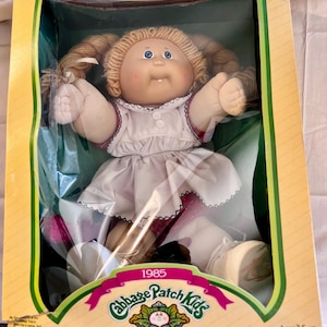 May include: A vintage Cabbage Patch Kids doll in its original box. The doll has blonde pigtails, blue eyes, and is wearing a white dress with red trim. The box features the Cabbage Patch Kids logo and the text "1985".