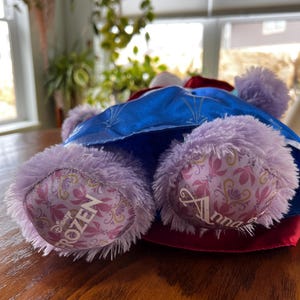 Puede incluir: Un peluche con un vestido azul con detalles de copos de nieve, una capa roja y un sombrero blanco. El juguete tiene patas lavanda con la palabra "Frozen" impresa en una pata y "Anna" en la otra.