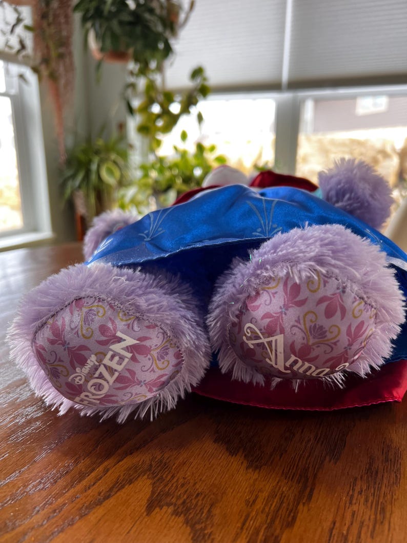 Puede incluir: Un peluche con un vestido azul con detalles de copos de nieve, una capa roja y un sombrero blanco. El juguete tiene patas lavanda con la palabra "Frozen" impresa en una pata y "Anna" en la otra.