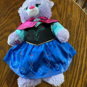 Puede incluir: Un gato de peluche morado que lleva un vestido azul con un corpi&ntilde;o negro y ribetes dorados. El vestido tiene un dise&ntilde;o floral. El gato tambi&eacute;n lleva una capa rosa y tiene ojos verdes y una nariz rosa. El juguete est&aacute; sobre una superficie de madera.