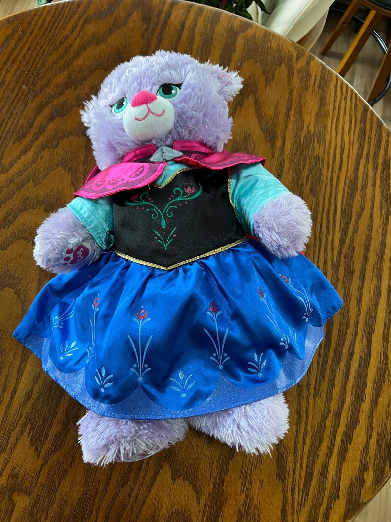 Puede incluir: Un gato de peluche morado que lleva un vestido azul con un corpi&ntilde;o negro y ribetes dorados. El vestido tiene un dise&ntilde;o floral. El gato tambi&eacute;n lleva una capa rosa y tiene ojos verdes y una nariz rosa. El juguete est&aacute; sobre una superficie de madera.