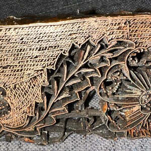 May include: A vintage wooden batik stamp with a floral design. The stamp is carved with intricate details, including flowers, leaves, and a geometric pattern. The stamp is made of dark wood and has a copper-colored finish.