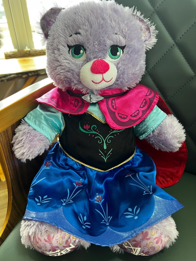 Puede incluir: Un oso de peluche lavanda que lleva un vestido azul y negro con detalles florales, una capa rosa y un forro de capa rojo. El oso tiene ojos verdes, una nariz rosa y est&aacute; sentado en una silla verde. Es un juguete para ni&ntilde;os.